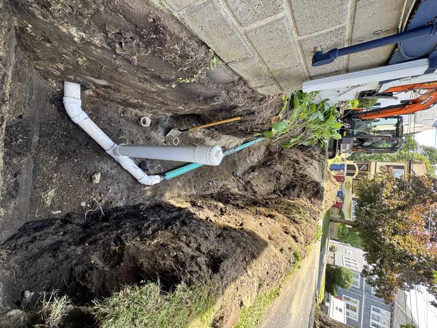 Open trench along a house foundation with a new sewer lateral and cleanout riser, excavator working at the far end